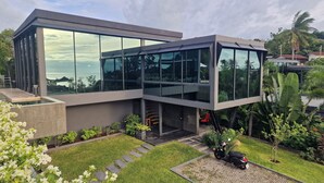 A modern two-story building with large glass windows, a concrete exterior, and a well-maintained garden with white flowers and a paved driveway. - Villa Obsidian (Koh Samui)