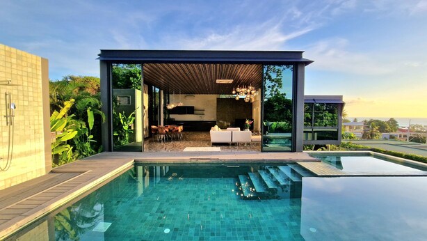 A modern outdoor pool area with a glass-enclosed lounge, a wooden pergola, and a view of the surrounding landscape. - Villa Obsidian (Koh Samui)