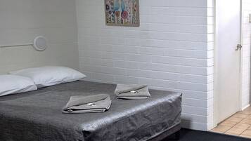 Ein Einzelbett mit grau strukturierter Matratze, weißen Kissen und einer gefalteten grauen Decke auf einem dunkelgrauen Teppich. Über dem Bett hängt ein farbenfrohes Blumenbild an der weißen Ziegelwand.