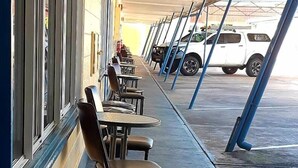 A covered outdoor seating area with tables and chairs, a row of parked vehicles, and a building with large windows.