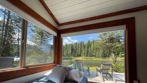 Un dormitorio con cama, almohadas y vistas a un lago y bosque a través de grandes ventanales.