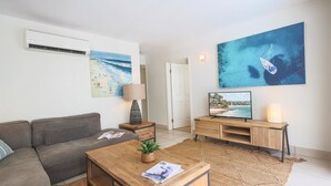A modern living room with a grey sofa, wooden coffee table, and a TV on a wooden stand. There are two paintings on the wall, one of a beach and another of a boat on the sea.