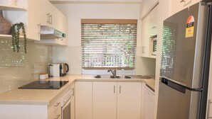 A modern kitchen with white cabinets, a stainless steel refrigerator, a built-in oven, and a window with blinds.