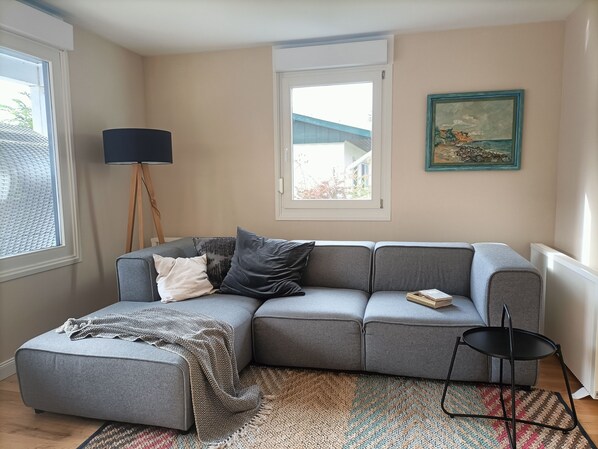 A living room with a grey sofa, a small ottoman, a black floor lamp, a patterned rug, and a painting on the wall.