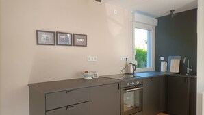 A modern kitchen with a built-in oven, a sink, and a window providing natural light. There are three framed pictures on the wall and a kettle on the counter. - Bungalow am Strand für die Ganze Familie (Insel Poel)
