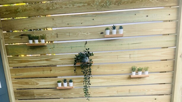 Un coin salon avec des fauteuils en osier et une petite table, un mur en bois orné d’étagères où sont disposées des plantes en pot, et un tapis rayé sur le sol.