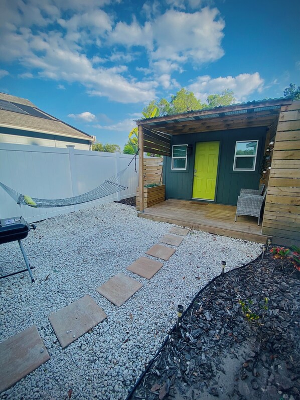 Una piccola struttura in legno con una porta verde, un portico e un patio in ghiaia con delle pietre.
