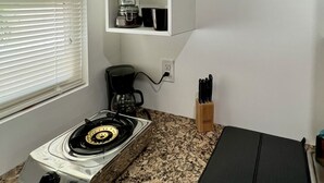 Une cuisine compacte équipée d’une cuisinière à gaz, d’une machine à café et d’un bloc à couteaux. Au-dessus du plan de travail, on trouve deux étagères : l’une supporte un mixeur, et l’autre contient un bocal et une tasse.