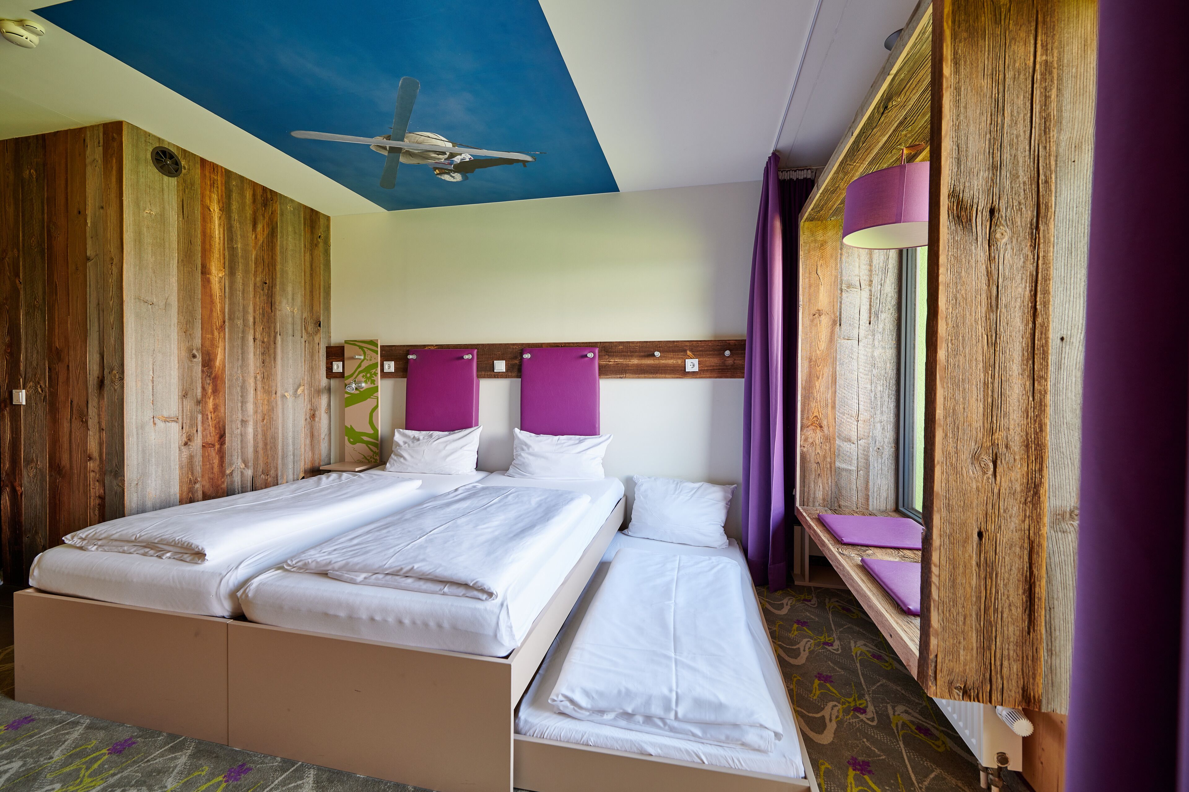 Ein Zimmer mit Holzwand, Deckenventilator, Bett mit weißer Bettwäsche und Fenster mit violetten Vorhängen.