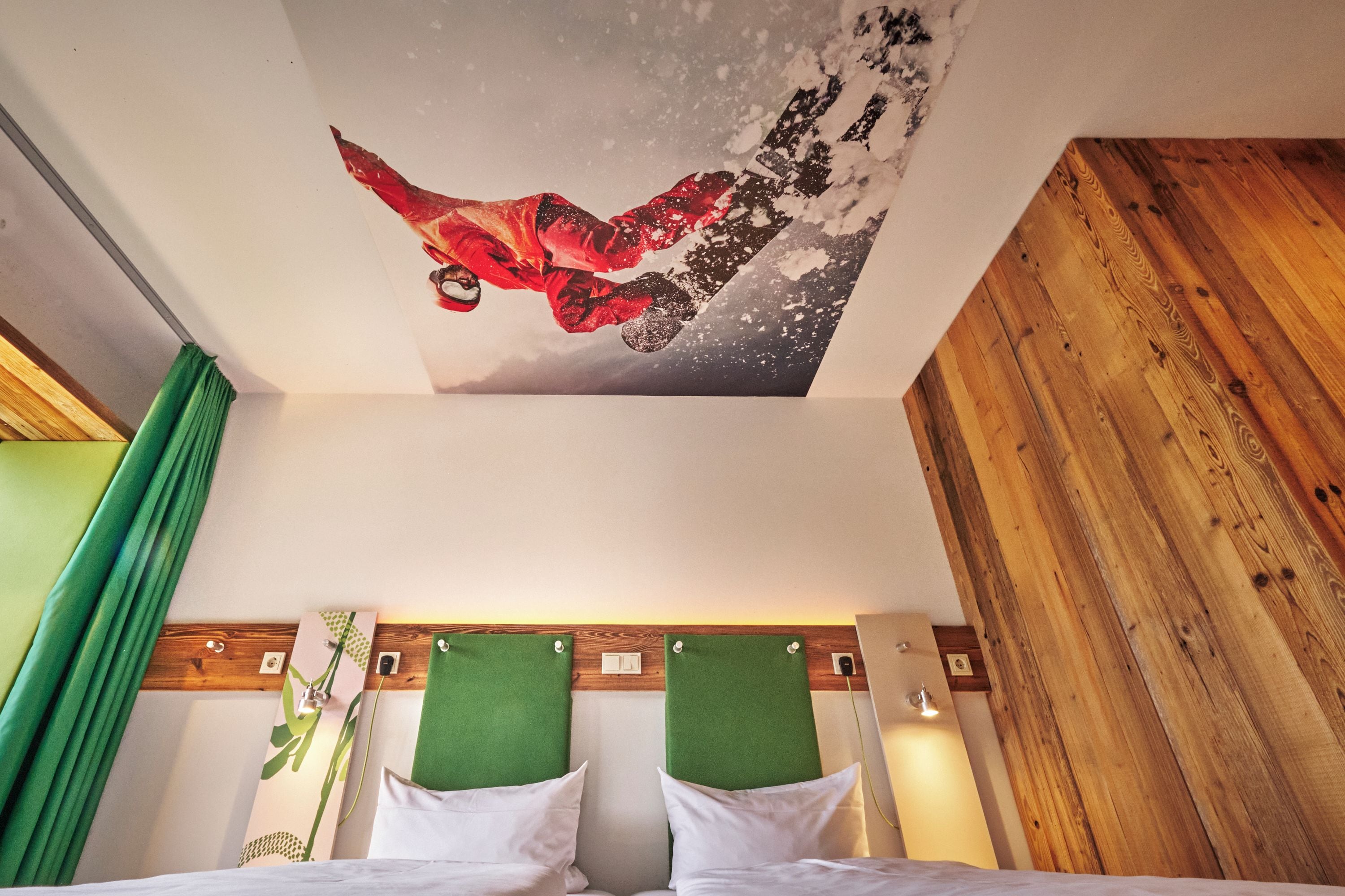 Una habitación de hotel con una cama, dos cabeceros verdes, una lámpara fijada a la pared y una obra de arte en el techo que representa a un snowboarder.