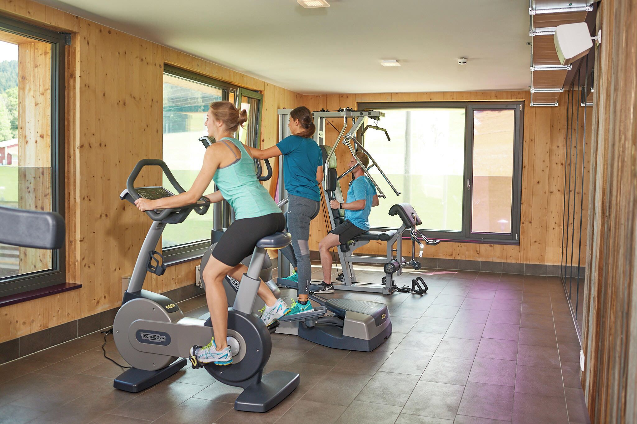 A gym with exercise equipment, large windows, and wooden walls.