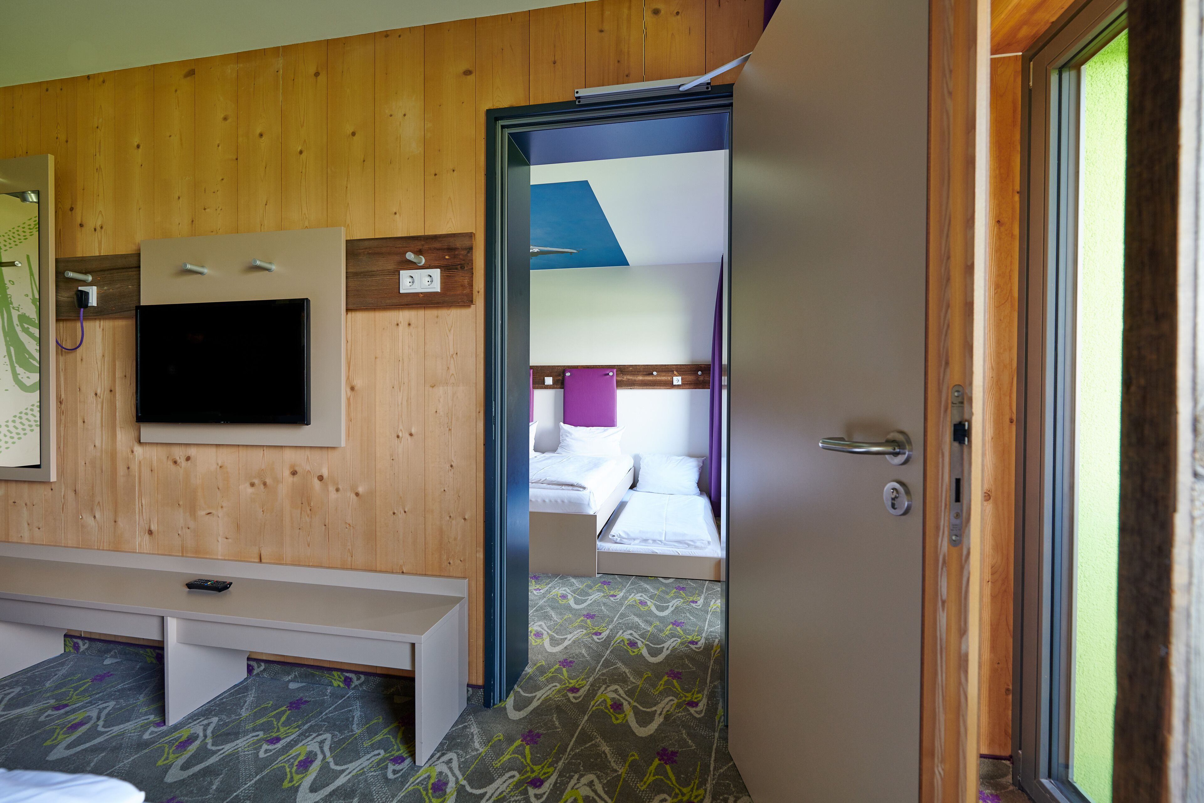 A hotel room with a wooden wall, a flat-screen TV mounted above a bed, a bench at the foot of the bed, and a door leading to another room.