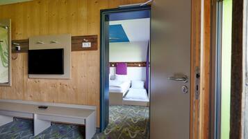 A hotel room with a wooden wall, a flat-screen TV mounted above a bed, a bench at the foot of the bed, and a door leading to another room.
