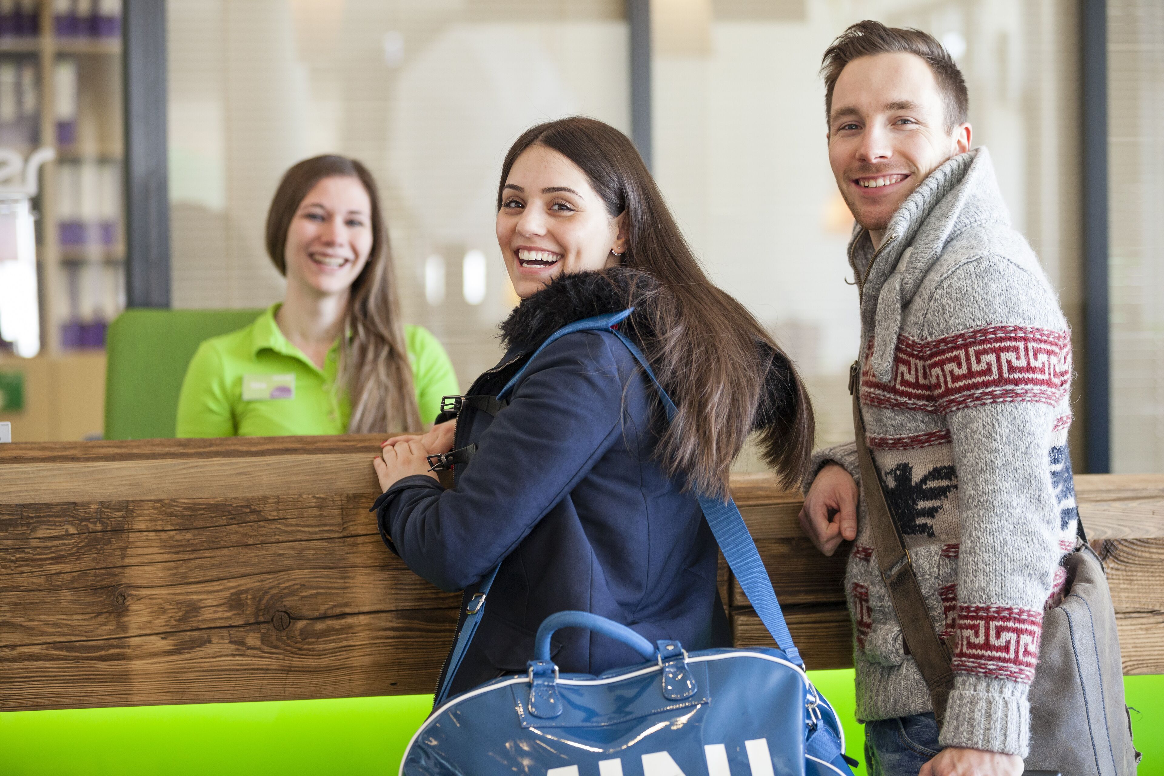 A reception area with wooden reception desk, green seating, and a person in a blue jacket.