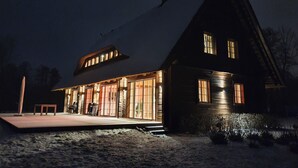Een groot, donker huis met een steil dak en een opvallende schoorsteen. Het heeft een goed verlichte veranda met een klein tafeltje en stoelen, en een zichtbare trap die naar de ingang leidt.