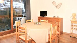 A dining area with a round table, wooden chairs, a television on a wooden cabinet, and a view of a snowy landscape through a large window. - 3-room apartment at the foot of the slopes with balcony (Le Corbier)