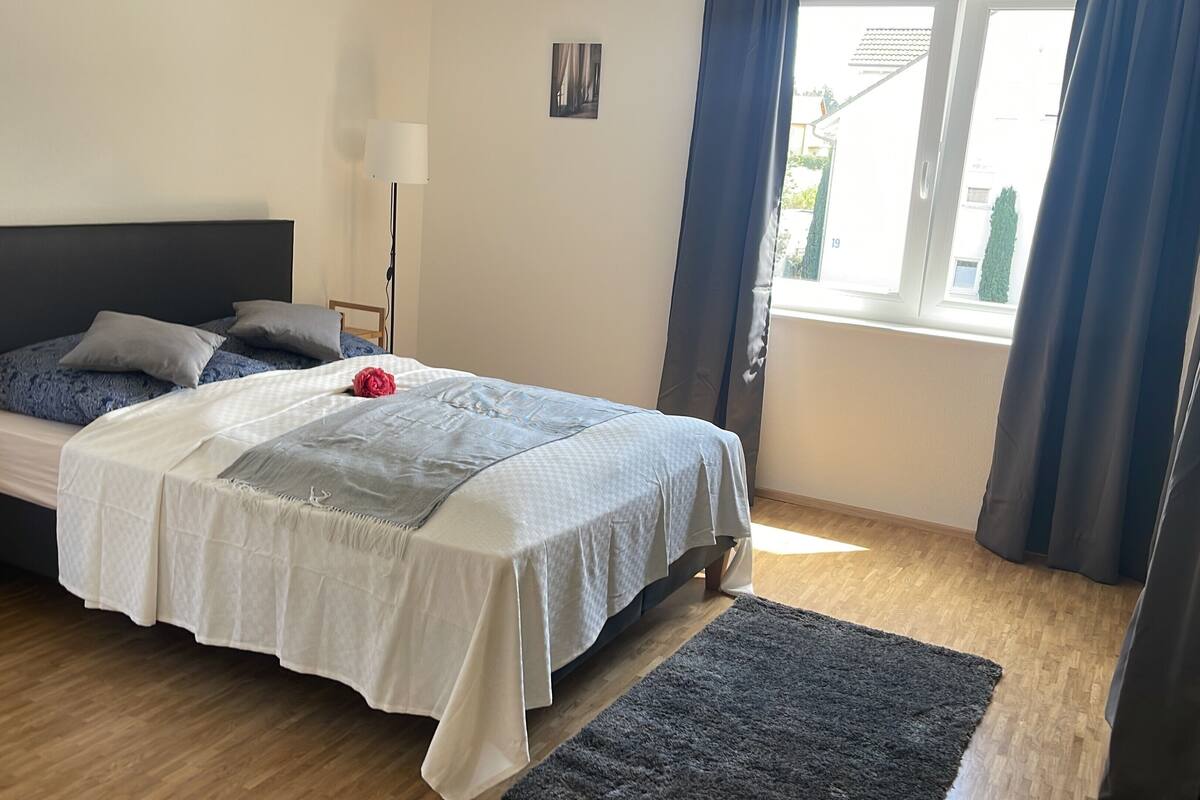 A bedroom with a bed, a nightstand, a lamp, a window with curtains, and a view of the outside.