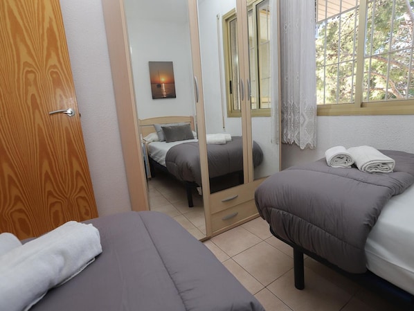 A room with two beds, a wooden door, a mirror, and a window with a view of trees. - Cozy 2BR Apartment with Pool, Wi-Fi, and Private Terrace in Salou's Lively Tourist Area (Salou)