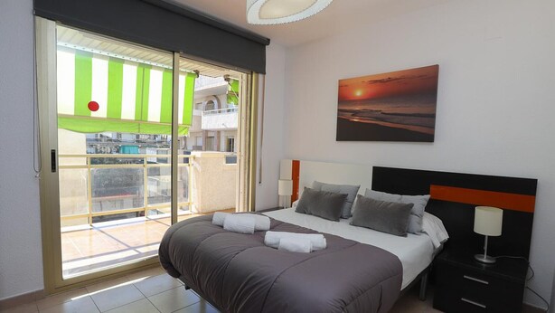 A bedroom with a bed, a nightstand, a lamp, a painting, and a sliding glass door leading to a balcony. - Cozy 2BR Apartment with Pool, Wi-Fi, and Private Terrace in Salou's Lively Tourist Area (Salou)