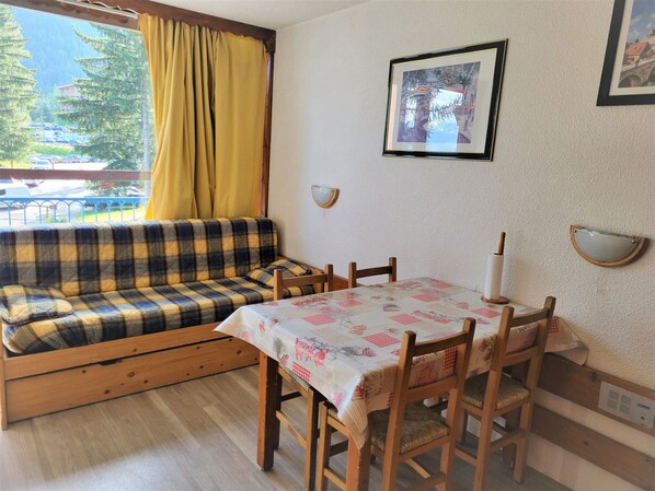 Quarto aconchegante com sofá, mesa de jantar e cadeiras. Há uma janela com cortinas, um quadro na parede e uma pequena prateleira fixada.