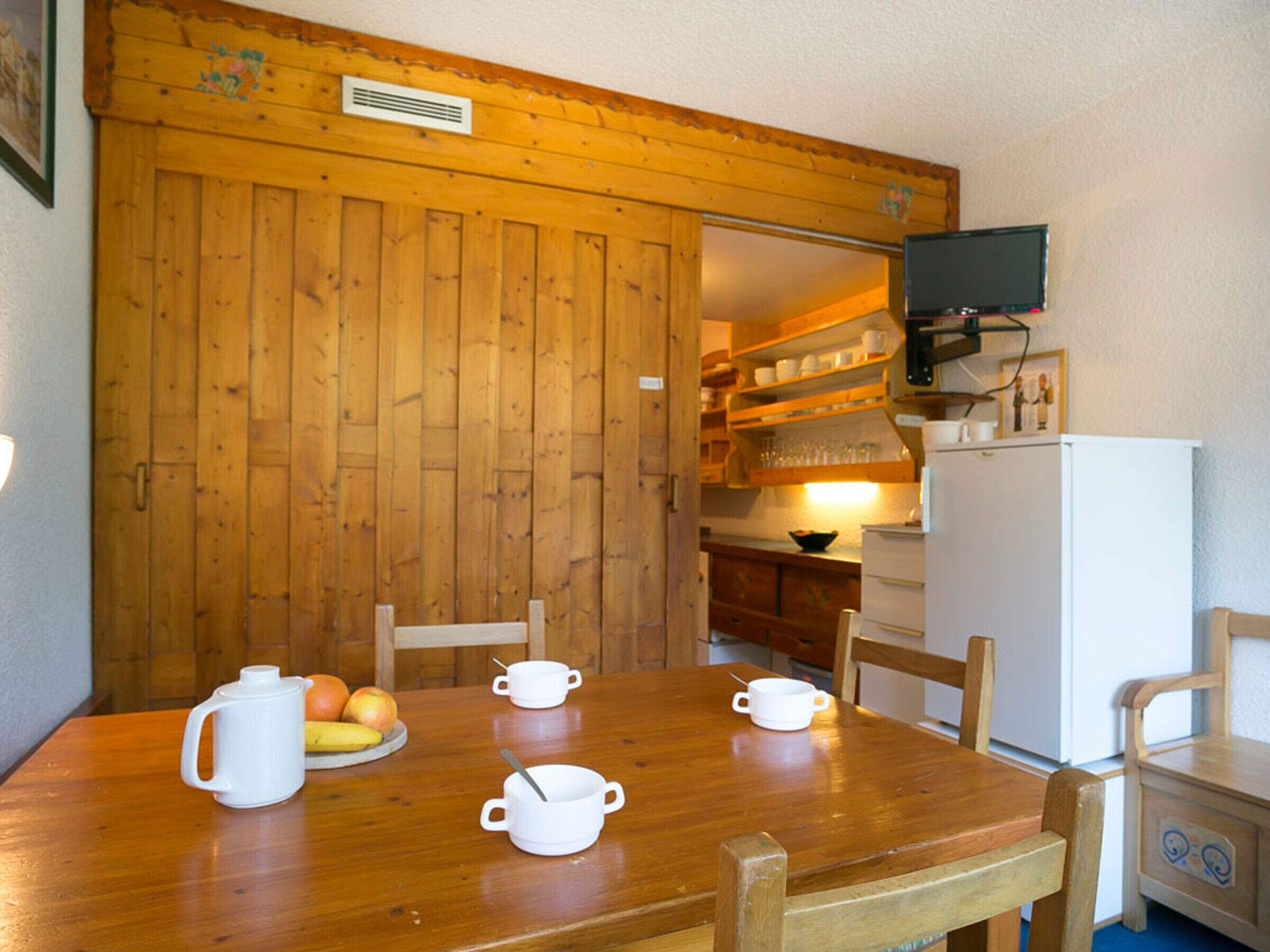 Une table de salle à manger en bois, avec des tasses blanches et un saladier de fruits. On aperçoit un réfrigérateur blanc et un téléviseur à l’arrière-plan.