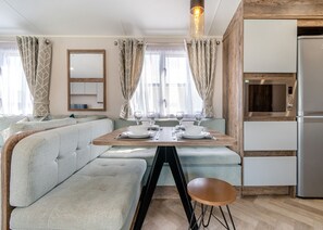 A modern kitchenette with a built-in microwave and refrigerator, a dining area with a wooden table and light-colored seating, and a large mirror above the table.