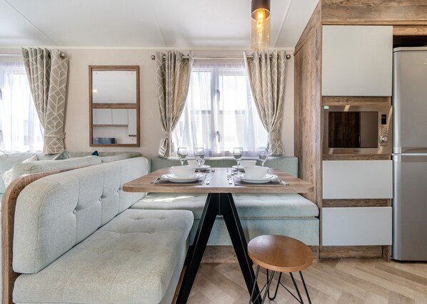 Una kitchenette moderna con microondas y refrigerador integrados, una zona de comedor con mesa de madera y asientos de tonos claros, y un espejo grande sobre la mesa.