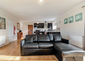 A living room with a black leather sofa, wooden flooring, a kitchen in the background, and a white ceiling.