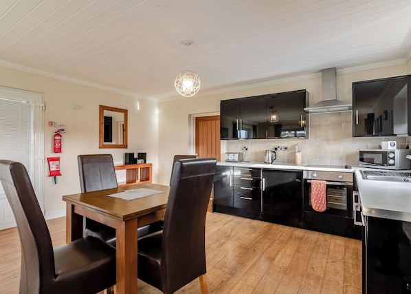A modern kitchen with black cabinets, a wooden dining table, and a hanging light fixture. - Luxury and contemporary retreat with a spacious open plan kitchen/living area. (Hornsea)