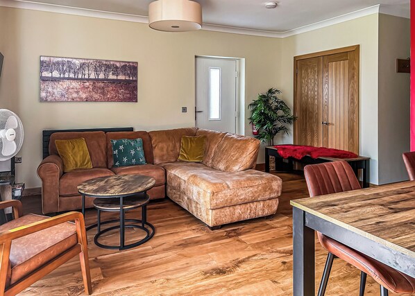 A living room with a sectional sofa, a round coffee table, a dining table with chairs, a fan, a painting, a door, and a wooden cabinet. - 3 bedroom accommodation in Flaxton (Flaxton)