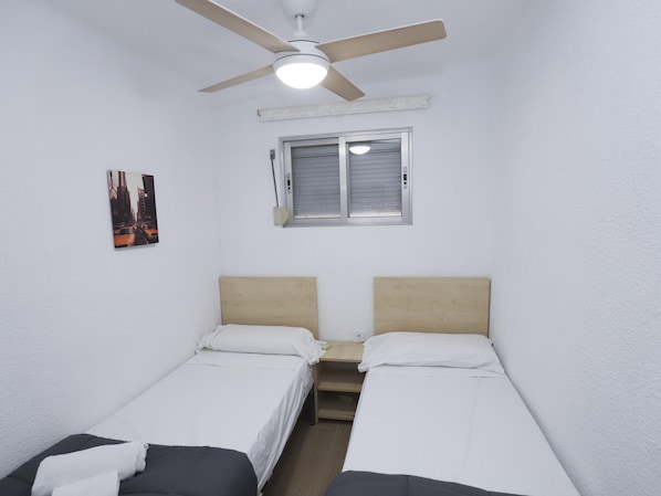 A room with two single beds, a ceiling fan, a window with blinds, and a wall-mounted picture.