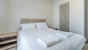 Una cama bien tendida con sábanas blancas y dos toallas dobladas, un cabecero de madera, una mesita de noche y una puerta a la derecha.