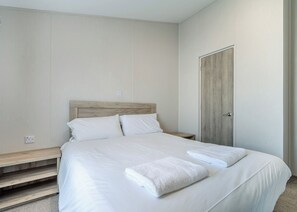 A neatly made bed with white linens and two folded towels, a wooden headboard, a bedside table, and a door to the right.