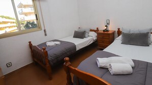 Habitación con dos camas, una silla de madera, una mesita de noche con lámpara y una ventana con vista a edificios.