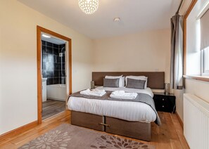 Un dormitorio con cama, mesitas de noche, ventana con cortinas, zona de ducha y puerta que da a otra habitación.