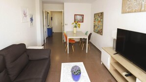 Una sala de estar con un sofá, una mesa de centro con una planta, un televisor sobre un mueble, un rincón de comedor con una mesa y sillas, y un pasillo que lleva a otra habitación.