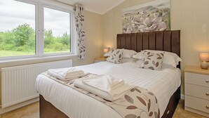 A bedroom with a large window, a bed with leaf-patterned bedding, a headboard, bedside lamps, and a painting on the wall. - Lakeview 3 Pet Friendly (Woodhall Spa)
