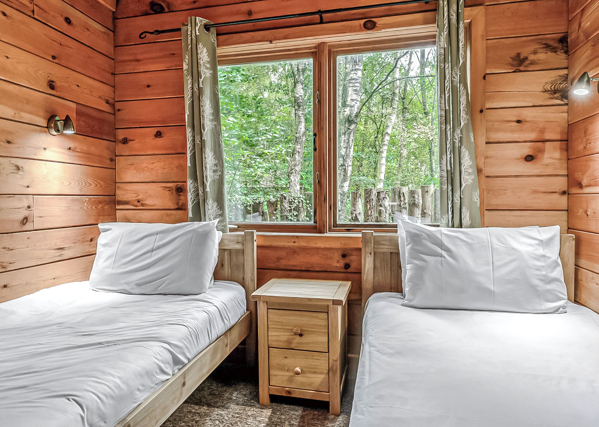 A cabin room with two beds, a small wooden nightstand, a window with curtains, and a view of trees.