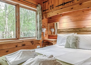 A wooden cabin bedroom with a bed, a nightstand, a mirror, and a window with curtains.