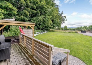 Une terrasse en bois avec des meubles d’extérieur, un espace salon couvert et une pelouse verdoyante offrant une vue sur les arbres et les collines.
