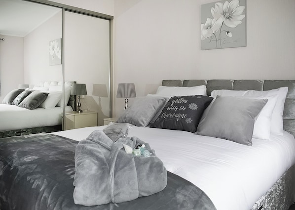 A bedroom with a bed, pillows, a blanket, a nightstand, a lamp, a mirror, and a wall art piece.