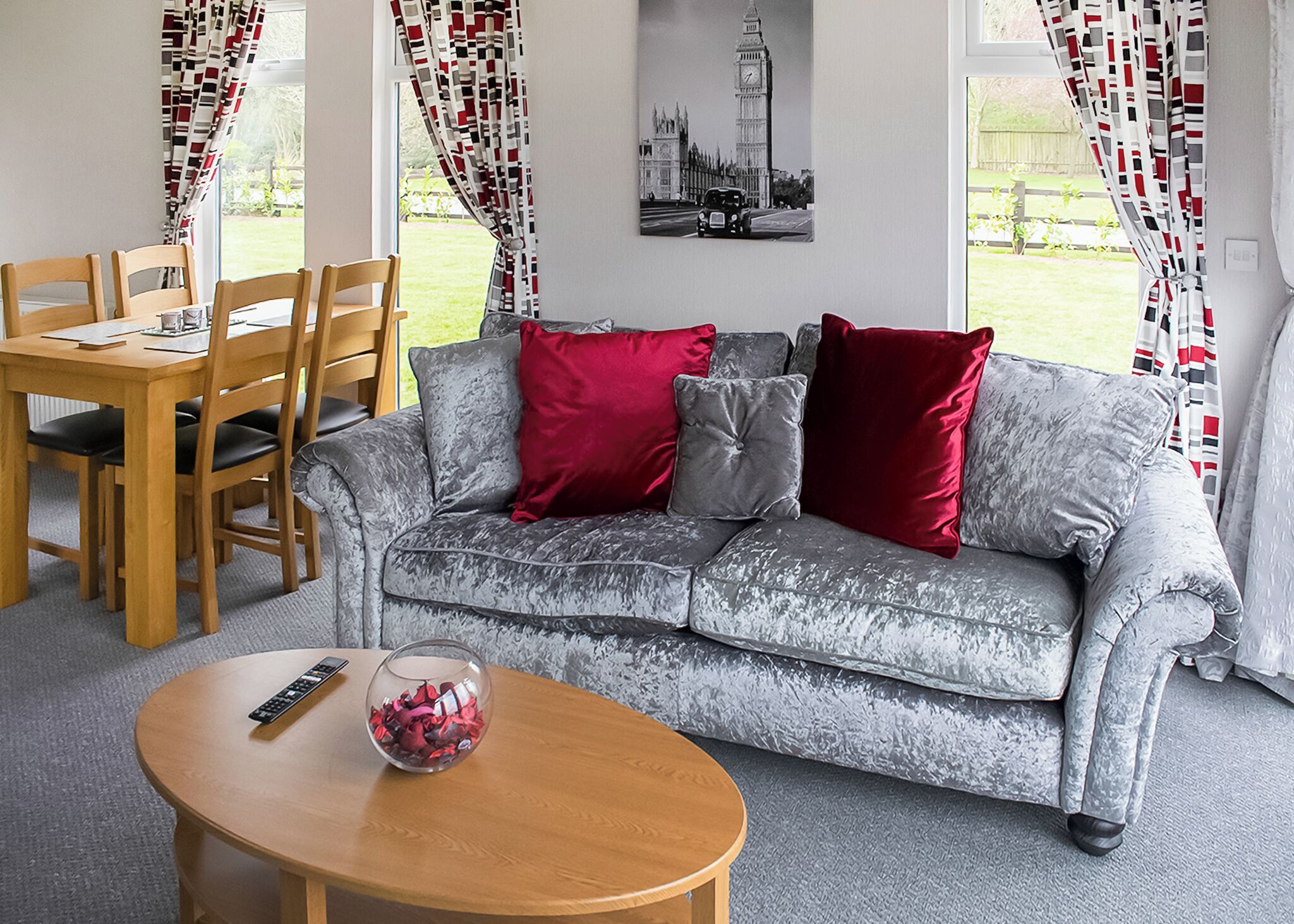 Una sala de estar con un sofá gris, cojines rojos y grises, una mesa de centro de madera con un control remoto, y un comedor con sillas y mesa de madera.