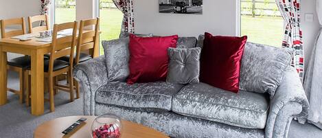 Una sala de estar con un sofá gris, cojines rojos y grises, una mesa de centro de madera con un control remoto, y un comedor con sillas y mesa de madera.