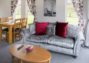 Un salon avec un canapé gris, des coussins rouges et gris, une table basse en bois avec une télécommande, et un coin repas avec des chaises et une table en bois.