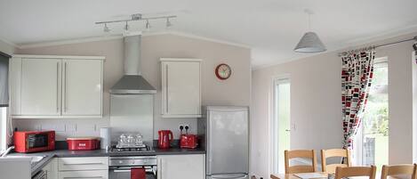 Une cuisine avec des meubles blancs, un micro-ondes rouge, une cuisinière à gaz, un réfrigérateur et un coin repas avec des chaises en bois.
