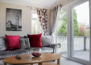 Un salon avec un canapé gris, des coussins rouges, un saladier en verre posé sur une table en bois et une grande fenêtre donnant sur un paysage verdoyant.