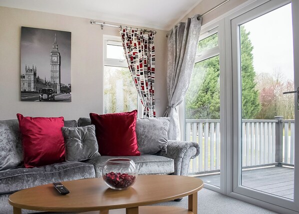 A living room with a grey sofa, red cushions, a glass bowl on a wooden table, and a large window with a view of greenery.