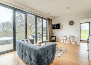 A modern living room with a grey sofa, a dining area with white chairs, a TV mounted on the wall, and a large window offering a view of trees and a hot tub outside. - Woodland View Retreat (Lewes)