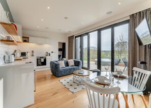 A modern kitchen with white cabinets, a blue sofa, and a dining area with a glass table and white chairs. - Woodland View Retreat (Lewes)