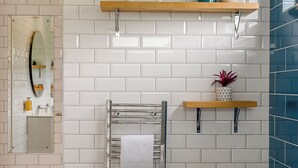 Une salle de bain avec des carreaux de céramique blancs rectangulaires, un porte-serviettes, un miroir et deux étagères ornées d’objets décoratifs.